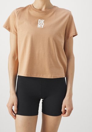 Persoon draagt een cropped lichtbruine DKNY T-shirt gecombineerd met zwarte geribbelde aansluitende shorts, tegen een effen achtergrond.