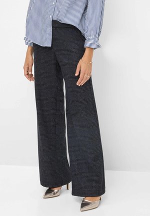 Femme portant un pantalon à carreaux foncés taille haute à jambes larges, une chemise rayée boutonnée et des talons pointus métalliques, debout devant un fond blanc.