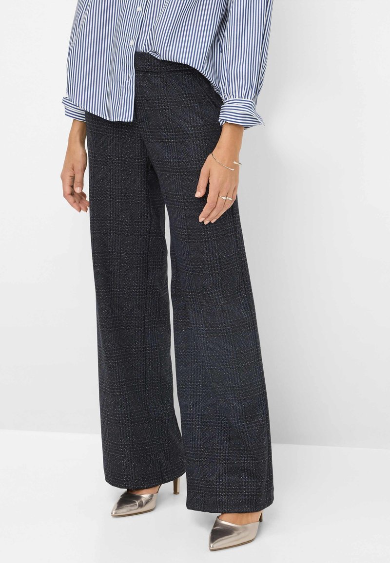 Femme portant un pantalon à carreaux foncés taille haute à jambes larges, une chemise rayée boutonnée et des talons pointus métalliques, debout devant un fond blanc.