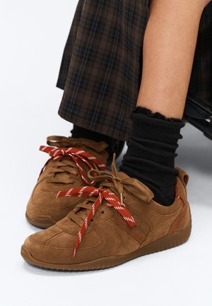 Baskets en daim marron avec des lacets orange et blancs, portées avec des chaussettes noires plissées et un pantalon à carreaux marron visible à la cheville.