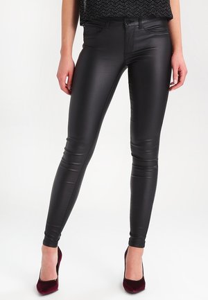 Nainen mustissa tekonahkaisissa kapeissa housuissa ja tummanpunaisissa samettikoroissa seisomassa yksinkertaisen valkoisen taustan edessä.