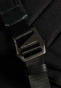 Tela negra con acabado texturizado que presenta una hebilla metálica y detalles de cuero. La hebilla está grabada con "Piquadro".