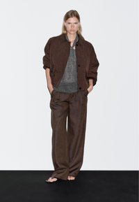 Frau steht mit Händen in den Taschen, trägt eine braune gesteppte Jacke, ein graues Strickoberteil, weite braune Hosen und schwarze Sandalen mit offenem Zehenbereich vor einem weißen Hintergrund.