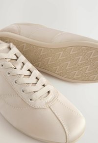 Beige sneakers with smooth leather upper, rounded shape, thick laces, padded collar, and textured rubber outsole featuring a zigzag pattern.