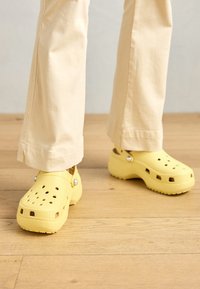 Yellow clogs made of lightweight material with ventilation holes. Features a rounded toe and a textured sole. Worn with light-colored trousers.