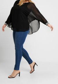 Blouse légère noire avec des manches transparentes et des poignets ornés de bijoux, portée avec un jean bleu slim et des sandales à talons noires. Tissu lisse et fluide.
