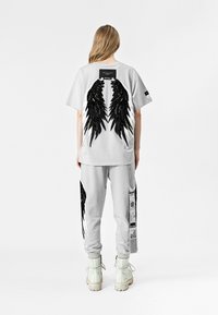 Camiseta gris de gran tamaño con gráficos de alas en negro, combinada con pantalones de chándal grises que tienen acentos de alas y detalles de etiqueta. Botas blancas.