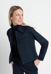 Marineblauwe jas met een kraag, voorzak, knoopsluiting en geribde manchetten. Glad textuur met contrasterend blauwe binnenkant.