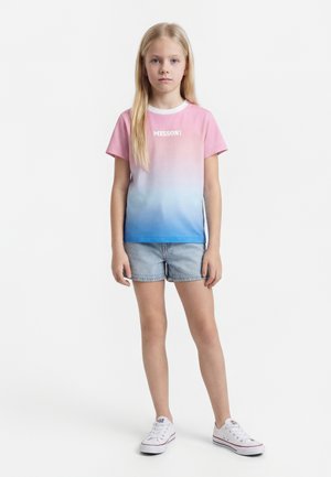 Ξανθιά κοπέλα φοράει ένα ροζ προς μπλε διαβαθμισμένο t-shirt Missoni, ανοιχτόχρωμα τζιν σορτς και άσπρα αθλητικά παπούτσια σε λευκό φόντο.