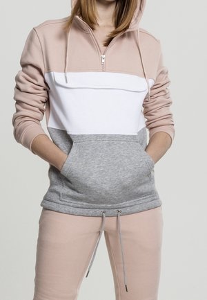 Personne portant un sweat à capuche color-block avec des sections beige, blanc et gris, mains dans la poche kangourou, associé à un pantalon beige.