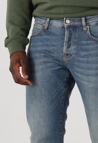 Jeans in denim di un azzurro chiaro con una finitura sbiadita, cinque tasche, cuciture visibili e un bottone metallico in vita. Mano appoggiata vicino.