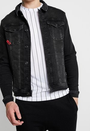 Man draagt een zwart denimjack over een wit overhemd met dunne verticale zwarte strepen, met één hand in de zak en de andere ontspannen.
