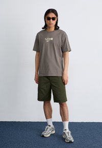 En ung mand står imod en simpel væg, iført solbriller, en grå t-shirt, grønne shorts og sneakers, med et selvsikkert udtryk.