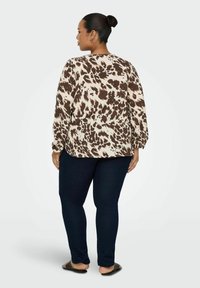 Majica s dolgimi rokavi v svetlo bež barvi z rjavimi abstraktnimi madeži, kombinirana z temno modrimi ozkimi kavbojkami. Očesne sandale so vidne. Gladka tekstura.