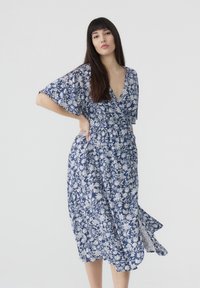 Abito a portafoglio floreale in blu e bianco, con maniche corte, scollatura a V e vita aderente. Presenta una gonna fluente all'altezza del ginocchio e un delicato motivo.