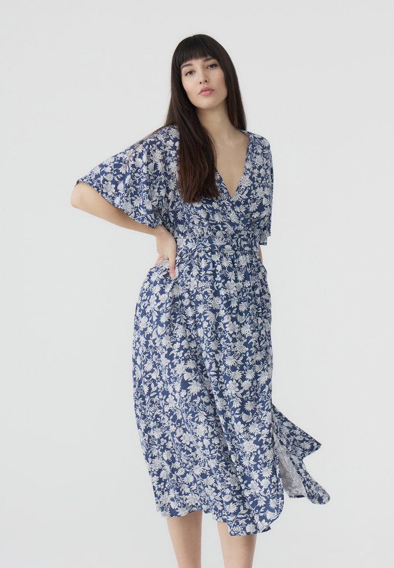 Abito a portafoglio floreale in blu e bianco, con maniche corte, scollatura a V e vita aderente. Presenta una gonna fluente all'altezza del ginocchio e un delicato motivo.