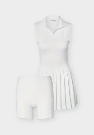 Bela brezrokavna polo obleka z gumbi spredaj in nabrano krilo, kombinirana z belimi oprijetimi kolesarskimi hlačami, na enobarvnem ozadju.