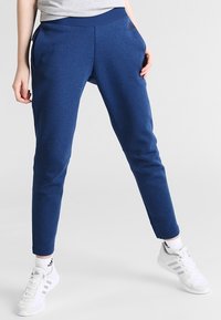 Osoba v modrých teplákových kalhotách Adidas, bílých teniskách Adidas a bílých ponožkách, stojící s rozkročenýma nohama na jednobarevném pozadí.