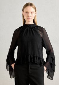Kvinna med rakt blont hår som bär stora hoopsmycken och en svart genomskinlig blus med långa ärmar, volanger i ärmsluten och nederkant, händerna i fickorna.