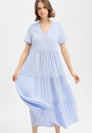 Frau trägt ein hellblau-weiß vertikal gestreiftes, kurzärmeliges V-Ausschnitt Stufenkleid in Midi-Länge und hält den Rock, während er sich nach außen ausbreitet.