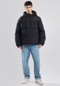 Black puffer jacket with a zip closure and a hood, featuring a quilted design. Worn over light blue jeans and white sneakers.
