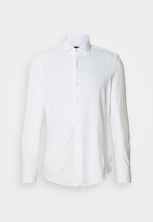 Garām piedurknēm balta krekla, izgatavota no mīksta auduma ar asu apkakli, dzeltenām pogām un noapaļotu apakšu. Pielāgots piegriezums ar aproču detaļām.