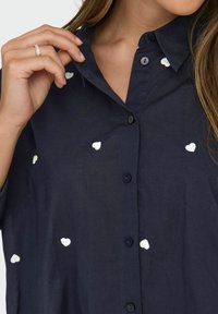 Button-up shirt in navy blue with short sleeves, featuring white embroidered heart shapes scattered across the fabric and small black buttons.