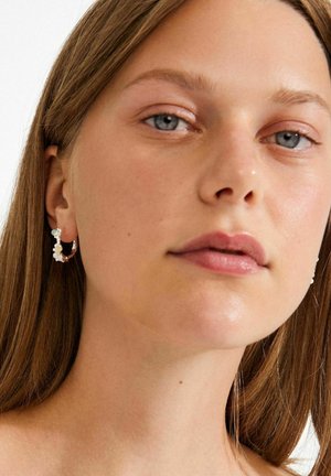 Zelta riņķa auskari ar nelielām caurspīdīgām pērlītēm un baltām ziedu detaļām. Modelim ir gari brūni mati un gluda āda.