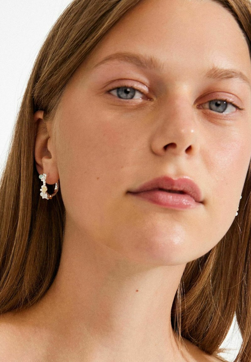 Gouden oorringen met kleine heldere kralen en witte bloemaccenten. Het model heeft lang bruin haar en een gladde huid.