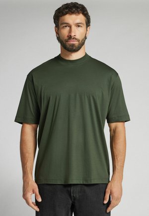 Bärtiger Mann mit kurzen lockigen Haaren trägt ein schlichtes dunkelgrünes kurzärmeliges T-Shirt und schwarze Hose, steht vor einem hellgrauen Hintergrund.
