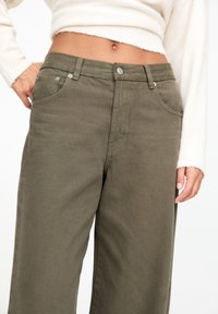 Pantalon large kaki en tissu de coton robuste, avec deux poches latérales et une fermeture à bouton. Un léger détail de couture ajoute de la texture.