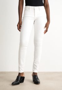 Person som bär vita slim fit-jeans och svarta klackskor, som står framför en plain vit bakgrund.