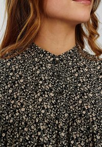 Blouse noire à fleurs avec col mandarin, petits boutons et taille froncée, ornée d'un motif délicat de fleurs blanches et roses.