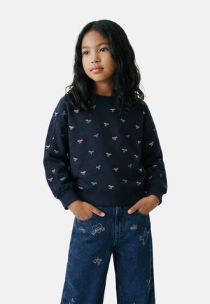 Jeune fille aux longs cheveux noirs portant un sweat-shirt bleu marine à petits motifs de nœuds et un jean bleu avec des broderies, les mains dans les poches.