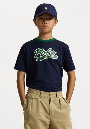 Mornarska pamučna majica s zelenim ovratnikom, bijeli i zeleni logo "Polo Ralph Lauren". Pješčane hlače s naborima i džepovima. Mornarska kapa s logotipom.