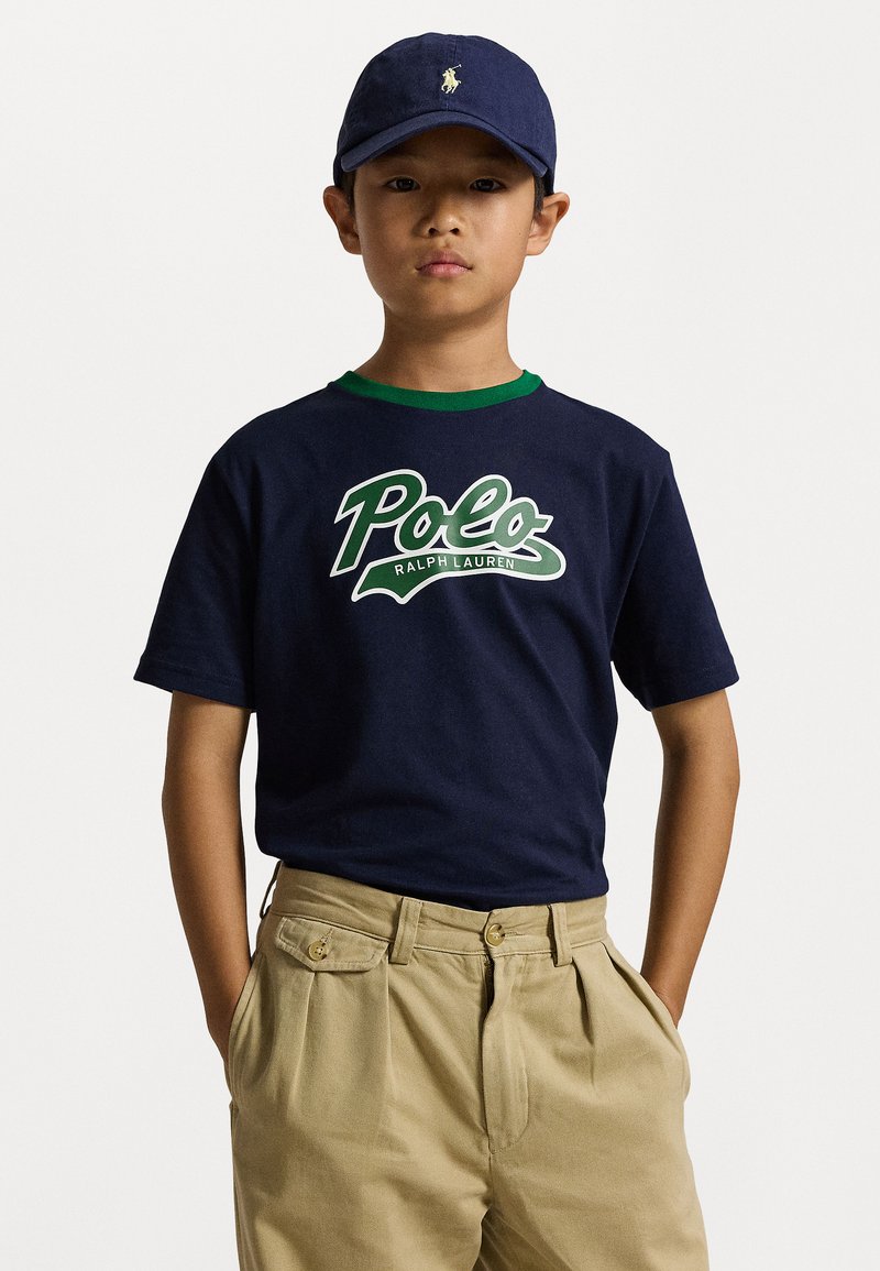 Mornarska bombažna majica z zelenim ovratnikom, belim in zelenim logotipom "Polo Ralph Lauren". Bež khaki hlače s pregibi in žepi. Mornarska kapa z logotipom.