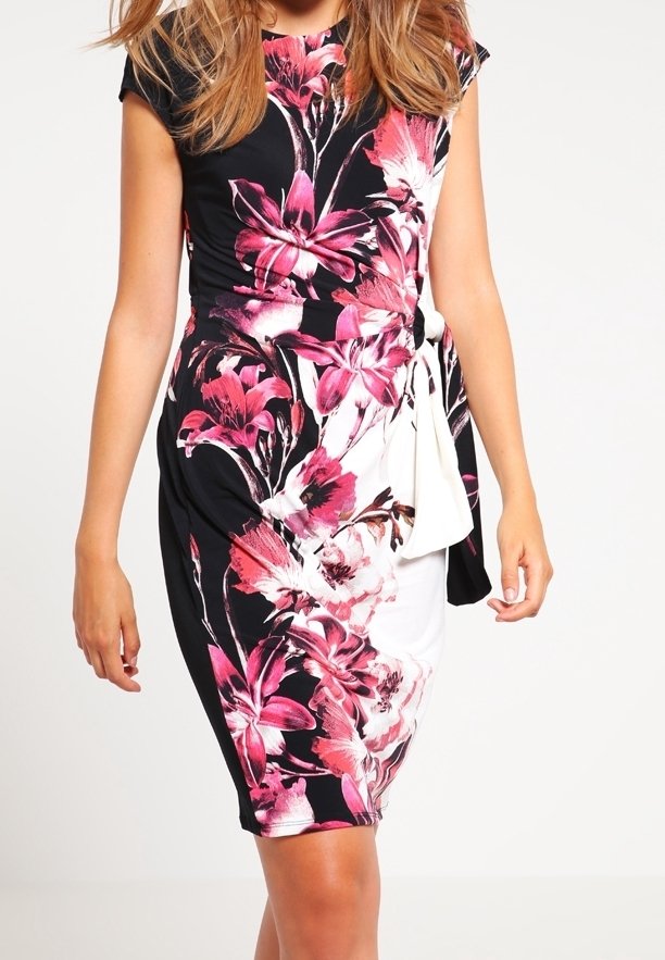 Femme portant une robe noire et blanche cintrée avec de grands motifs floraux roses et un détail de nouage à la taille, debout devant un fond uni.