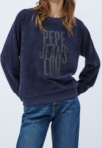 Femme portant un sweat-shirt bleu marine avec le texte "Pepe Jeans London" et un jean bleu, debout les mains derrière le dos.