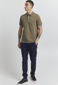 Oliven grøn polo-shirt med krave og tre knapper, parret med marineblå bukser og sorte sneakers. Viser synlige tatoveringer på armene.