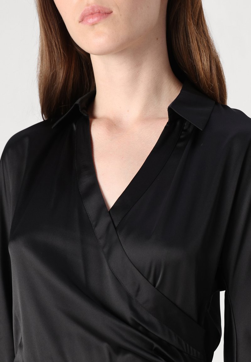 Schwarze Satinbluse mit Wickeldesign, die über einen Spitzkragen und eine glatte, glänzende Textur verfügt. Zu den Details gehört ein tiefes V-Ausschnitt.