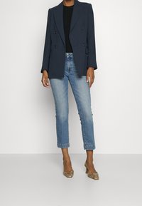 Osoba v námořnickém dvouřadovém saku, černém topu, světlých modrých zkrácených džínách a vysokých podpatc ích se vzorem hadí kůže stojící.