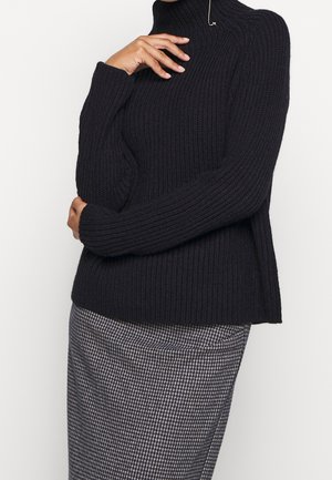 Persoon draagt een zwarte geribbelde coltrui en een zwart-witte houndstooth-rok tegen een effen achtergrond.