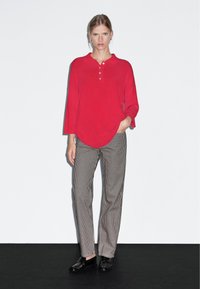 Polo shirt rosso a maniche lunghe con colletto e bottoni, abbinato a pantaloni a righe verticali in bianco e nero. Modello che indossa scarpe nere.