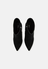 Bottines noires à bout pointu, fabriquées en matériau souple, avec une fermeture éclair sur le côté et une texture lisse. Logo visible à l'intérieur.