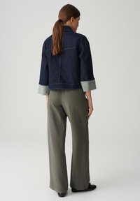 Giacca di denim scuro con un design cropped e polsini in denim chiaro, abbinata a pantaloni larghi verde oliva, vista da dietro.