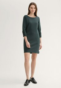 Sötétzöld, textúrált, hosszú ujjú ruha, csónak nyakkivágással, kissé térd felett, fekete fűzős cipőkkel párosítva.