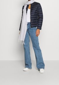 Person wearing navy quilted jacket, white tank top, light blue wide-leg jeans, white sneakers, and long light gray scarf.