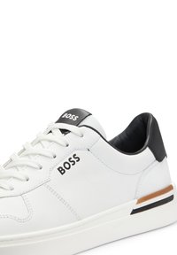 Vita lädersneakers med svart hälaccent, platta vita snören och svart "BOSS"-logotyp på sidan. Har ett subtilt brunt och svart rand på sulan.
