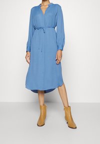 Donna che indossa un vestito midi blu a maniche lunghe con vita regolabile e stivali alla caviglia beige, in piedi contro uno sfondo chiaro e semplice.