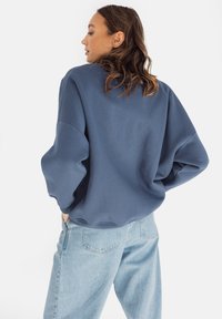 Niebieska bluza z obniżonymi ramionami i luźnym krojem, zestawiona z jasnoniebieskimi dżinsami. Materiał wydaje się miękki o gładkiej fakturze.